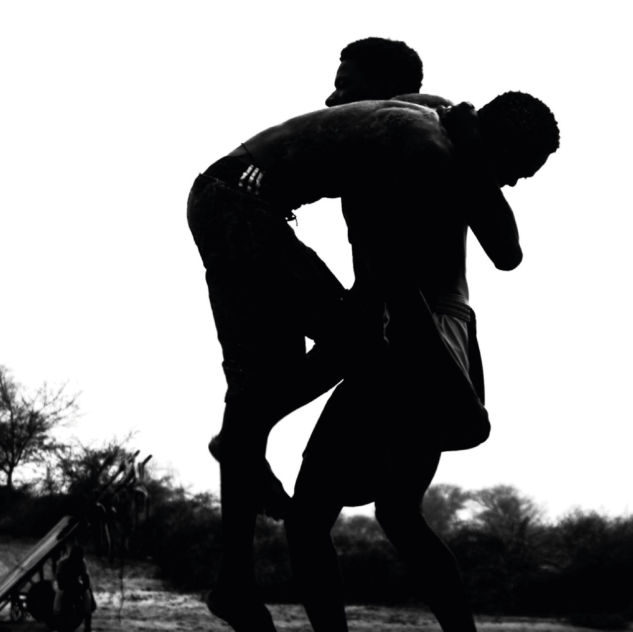 Dynamic black and white silhouette of two men wrestling in an outdoor setting, showcasing strength.